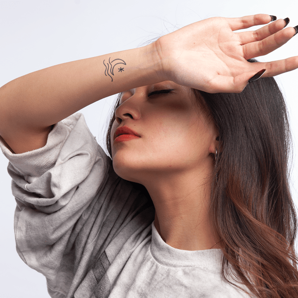 Người đang che mặt bằng tay, trên cổ tay có hình xăm nhỏ, nền trắng.