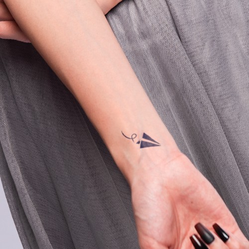 Cánh tay người với hình xăm máy bay giấy nhỏ trên cổ tay, mặc váy xám, móng tay sơn đen.