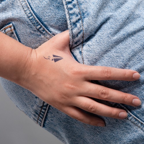 Bàn tay đặt trên túi sau của quần jean, có hình xăm chiếc máy bay giấy trên mu bàn tay.