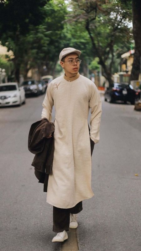 Người đàn ông mặc áo dài màu kem, đội mũ, đi giữa phố, xung quanh là xe cộ và cây cối.